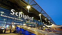 Illuminated entrance of Amsterdam Schiphol Airport at dusk, showing glass facade and signage.